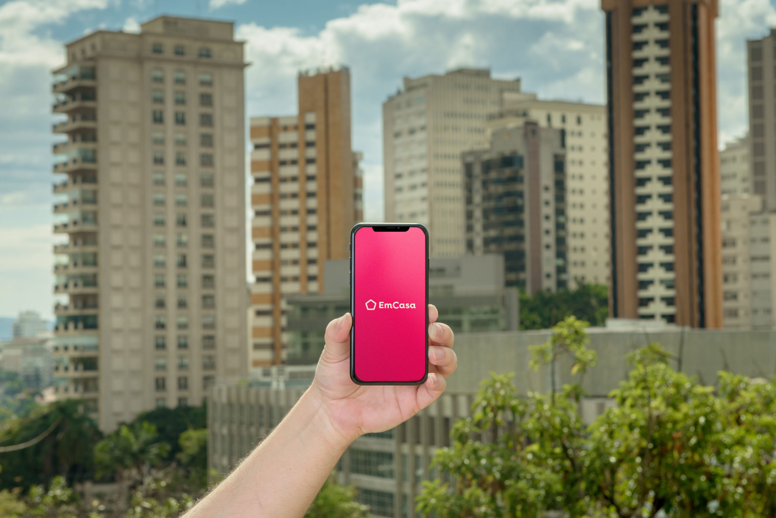 A imagem mostra uma pessoa segurando um celular com o logo da EmCasa, em um fundo cheio de prédios