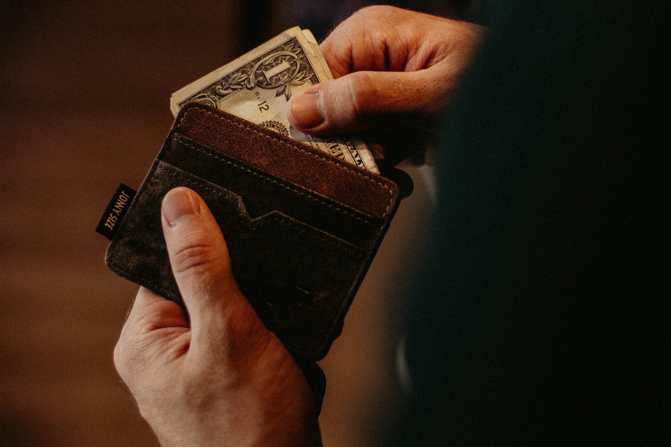 A imagem mostra uma carteira de couro marrom com uma nota sendo puxada pelas mãos de um homem, ilustrando o tema do texto
