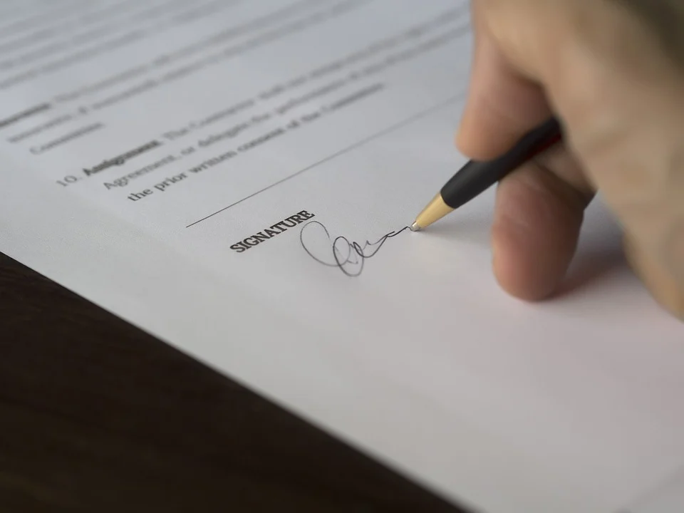 Um documento com palavras em inglês e a mão de uma pessoa branca utilizando uma caneta para redigir a assinatura no campo destinado a esse objetivo.