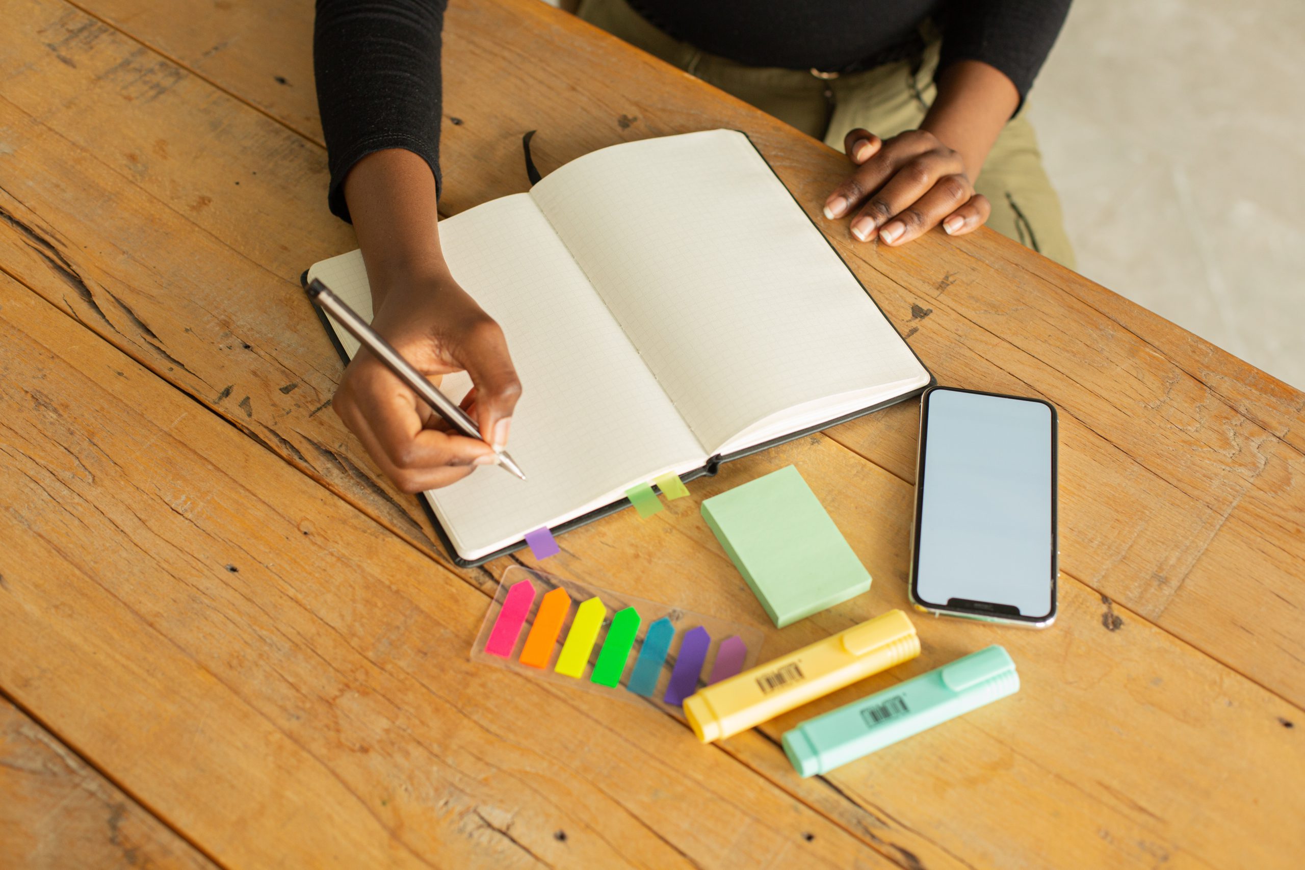 Em cima de uma mesa de madeira, há um caderno com folhas em branco, pequenos blocos de notas coloridos, canetas marcadoras azul e amarela, e um celular. As mãos de uma mulher negra seguram uma caneta prestes a escrever algo no caderno.